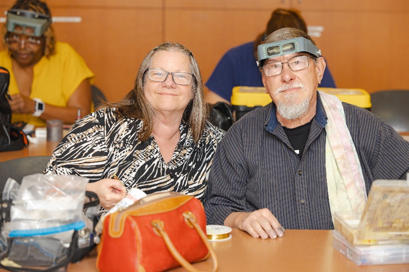 Nick and Deborah Carpenter sit together in an Allan Hancock College Community Education art class, smiling at the camera. Art supplies and project materials are spread across the table in front of them, while other students work in the background, reflecting the couple’s shared tradition of taking community art classes together.